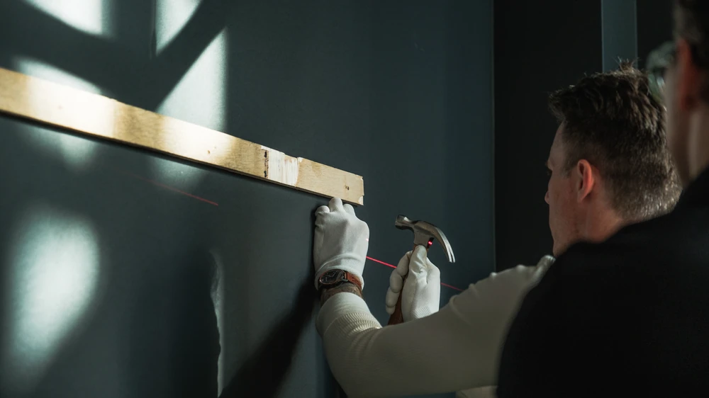 An installer in white gloves using a hammer beside a laser line and patterned window light.