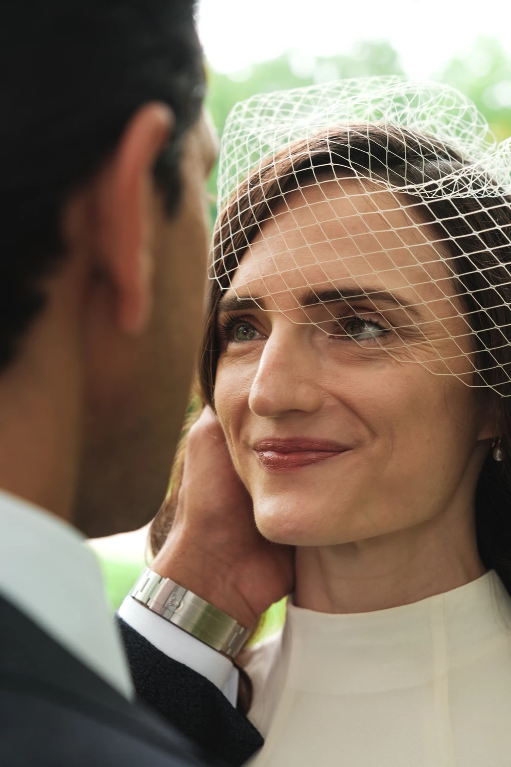A bride looking toward her partner through a delicate veil as a hand rests on her cheek.